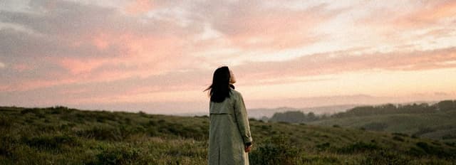 A woman stands on a hill and looks up at a wide pink evening sky